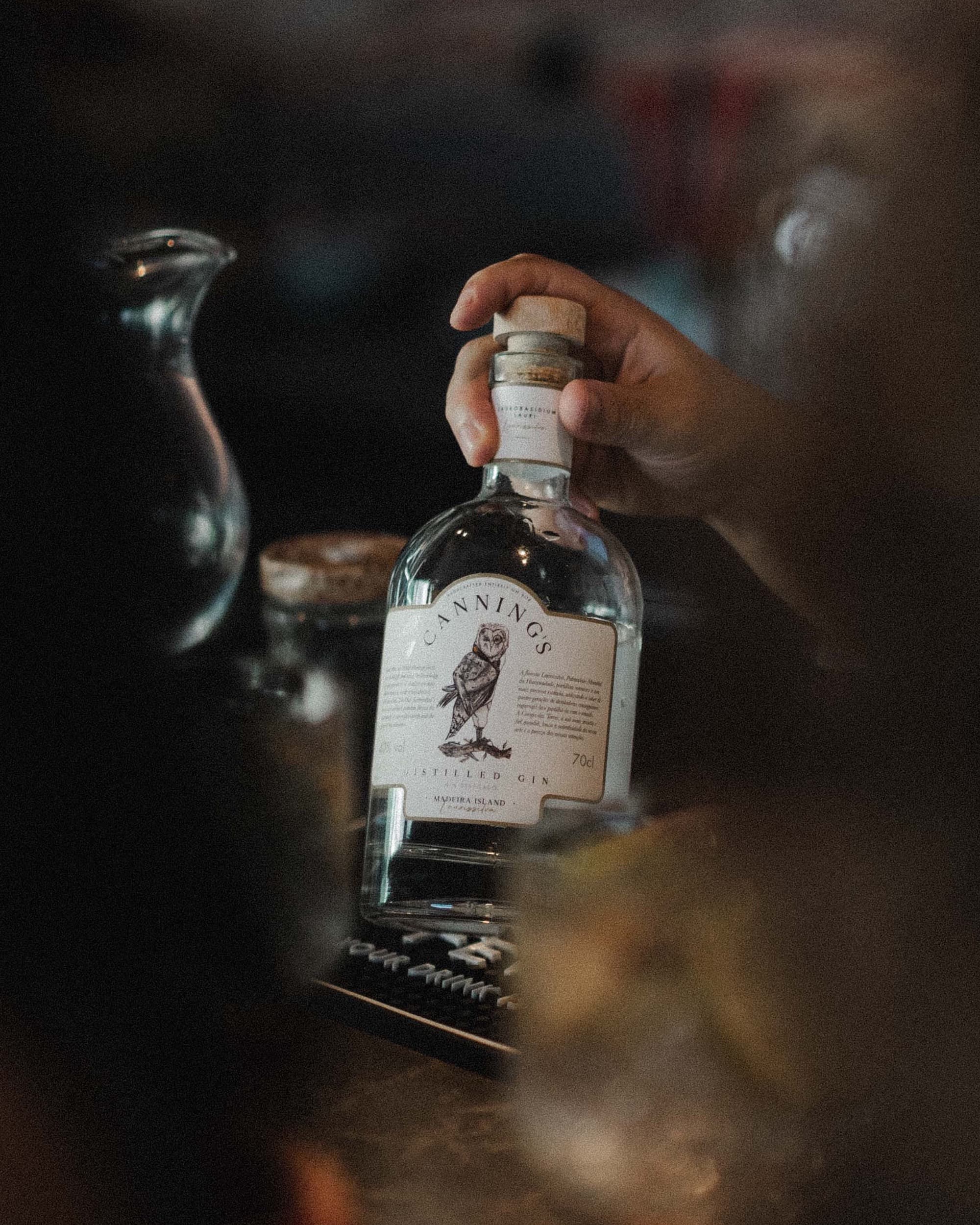 A hand lifting a bottle of Canning's gin by its cork, at a bar in warm light