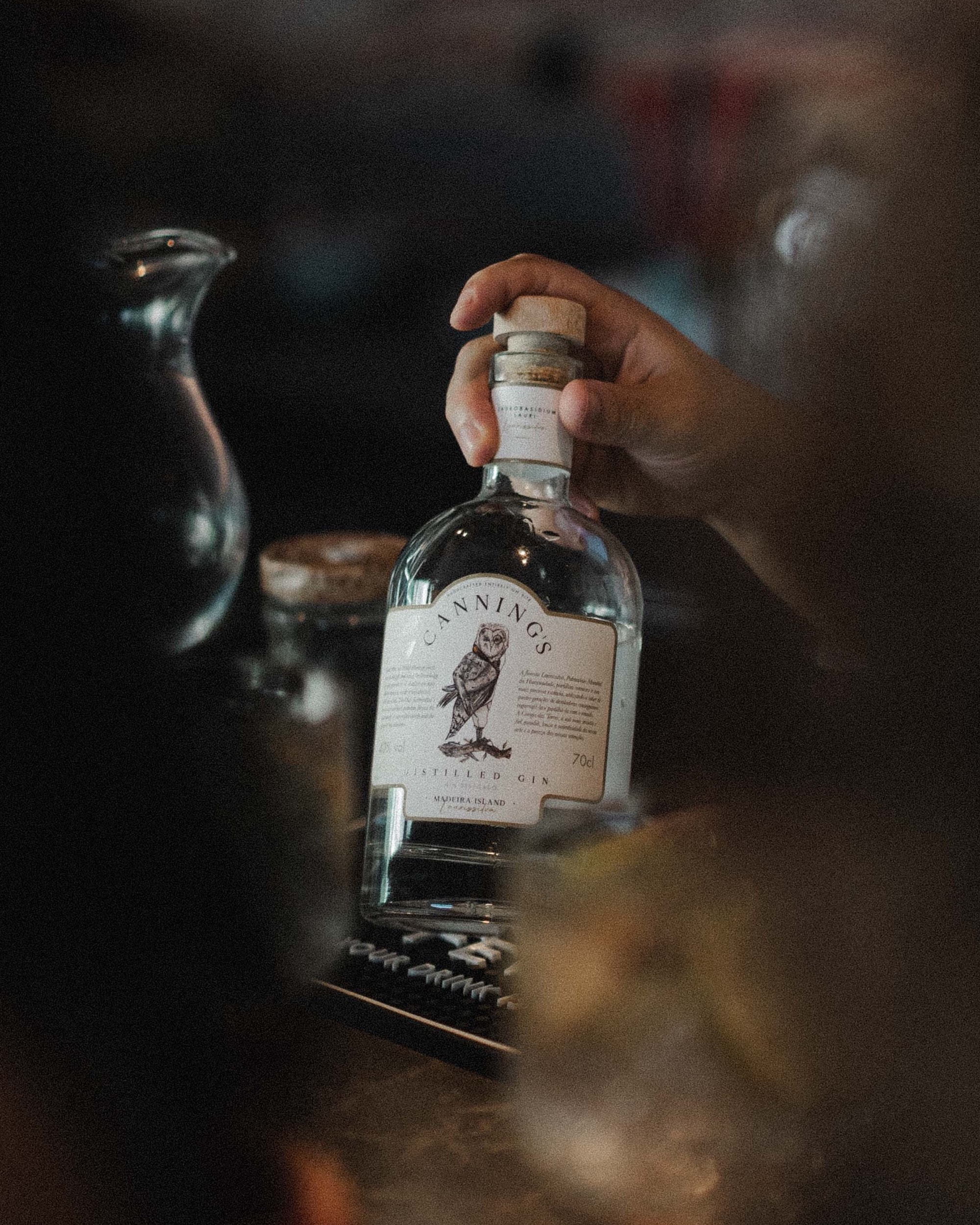A hand lifting a bottle of Canning's gin by its cork, at a bar in warm light