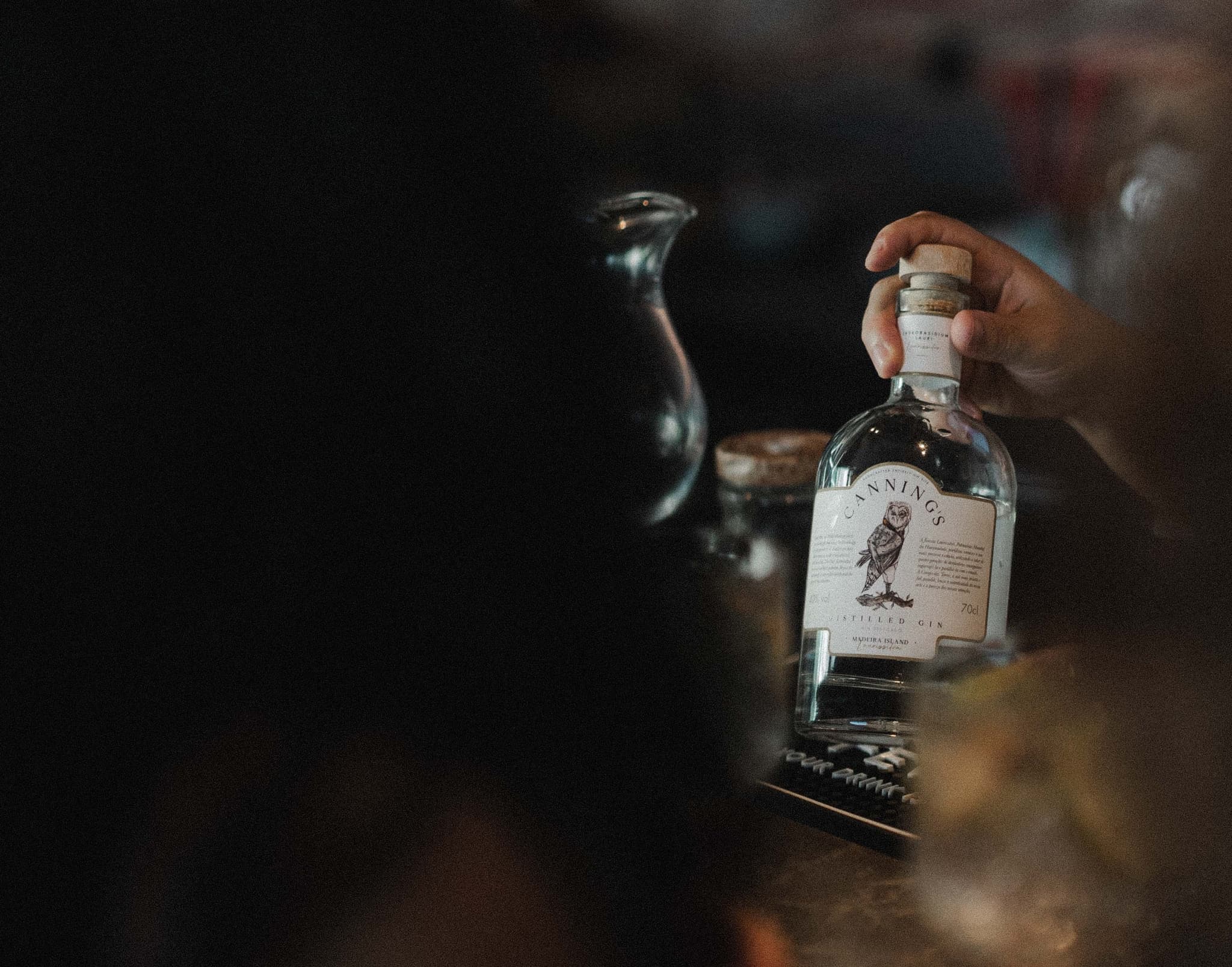 A hand lifting a bottle of Canning's gin by its cork, at a bar in warm light
